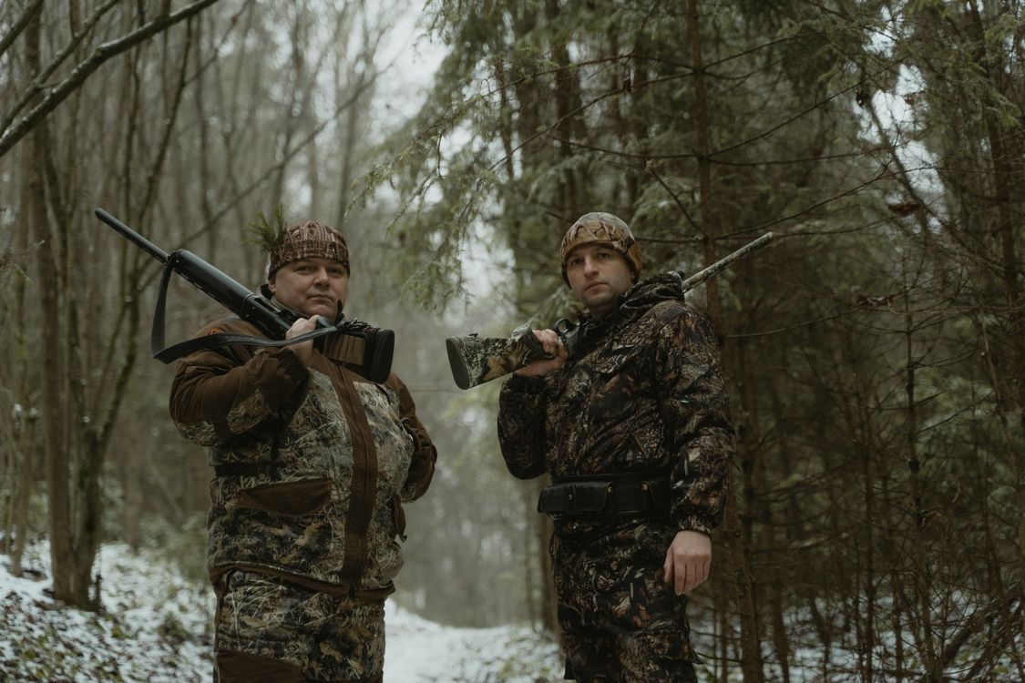 Two hunters in camouflage with rifles in snowy forest - deer hunting